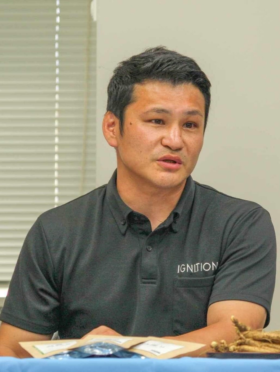 佐藤吏さん。IGNITIONのポロシャツ姿で、机に置いた高麗人参の前で話しているところ。